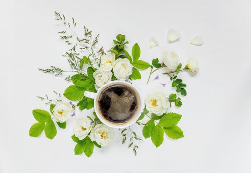 Decorative composition in retro style consisting of black coffee in a white porcelain cup and white wild rose flowers with green leaves on white background. Top view, flat lay