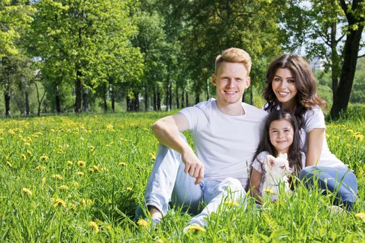 Portrait of happy smiling family of parents and girl sitting on grass with dandelion flowers at sunny summer day