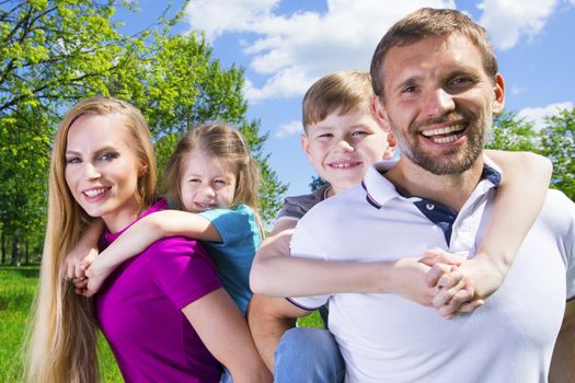 Happy family portrat, couple giving two young children piggyback rides smiling in summer park