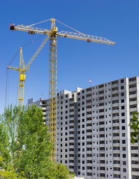 Two different tower cranes with latticed booms on a construction of a multi-story residential building and trees in the foreground 
