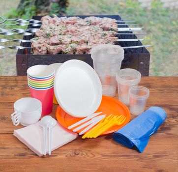 Different disposable plastic and paper cutlery, paper napkins and roll of disposable garbage bags on the old wooden planks against the background of barbecue grill with grilled skewered meat outdoors

