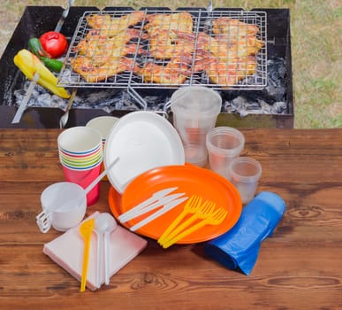 Different disposable plastic and paper cutlery, paper napkins and roll of disposable garbage bags on the old wooden planks against the background of charcoal grill with grilled skewered meat outdoors
