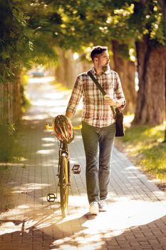 Handsome businessman going to work by bicycle. He is walking with coffee to go next to bike and thinking.
