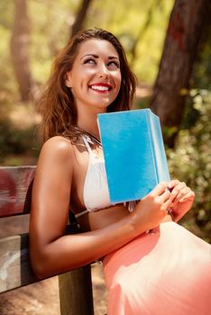 Young woman reading a book while relaxing in the forest and looking away with smile on her thinking face.