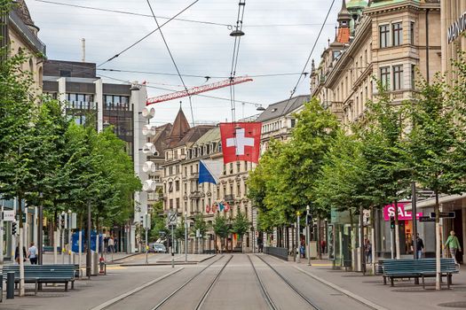 Zurich, Switzerland - June 10, 2017: Shopping promenade Bahnhofstrasse, inner city of Zurich. Swiss flag and tram railroad tracks in front.