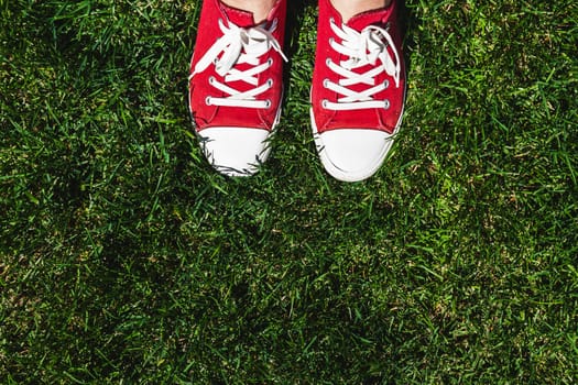 Legs in old red sneakers on green grass. View from above. The concept of youth, spring and freedom.