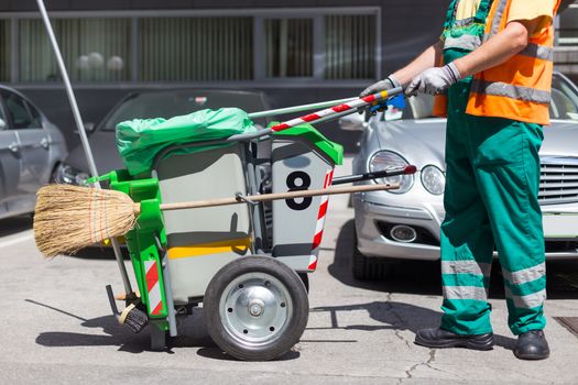 Worker of cleaning company in green uniform with garbage bin and a broom. Utility service company men worker of municipality sweeping city streets.