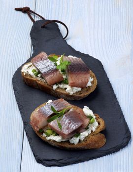 Delicious Sandwiches with Marinated Herring, Cream Cheese and Chives on Slices of Brown Bread closeup on Slate Board on Wooden background