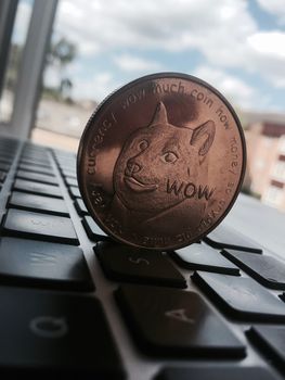 Digital currency physical brass dogecoin coin on the black computer keyboard near window.