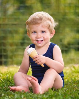 Sweet toddler baby boy eighteen months old, eating tasty ice cream over background with green bokeh outdoor.