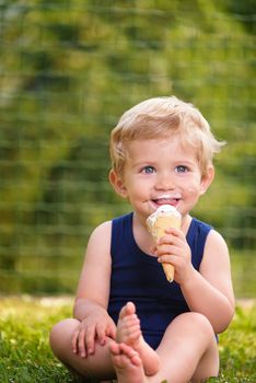 Sweet toddler baby boy eighteen months old, eating tasty ice cream over background with green bokeh outdoor.