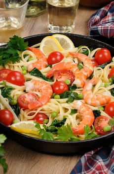 Pasta with fried prawns, peas, tomatoes and spinach in a frying pan, on a table with cider glasses.