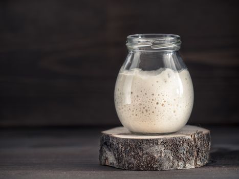 Active wheat sourdough starter in glass jar on brown wooden background. Starter for sourdough bread. Toned image. Copy space.