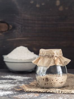 Active rye sourdough starter in glass jar and rye flour on brown wooden background. Starter for sourdough bread. Toned image. Copy space. Vertical