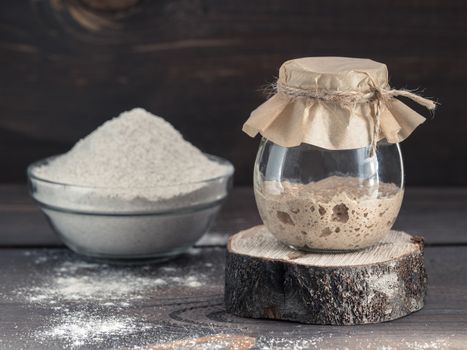 Active rye sourdough starter in glass jar and rye flour on brown wooden background. Starter for sourdough bread. Toned image. Copy space.