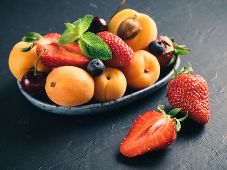 Closeup view of fruits and berries on dark background. Heap of fresh strawberries, blueberries, apricot and mint leaves in trendy plate. Focus on strawberry. Healthy food,superfood,diet,detox concept.