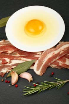 Raw pork bacon slices, rashers, egg yolk in white saucer, spices and peppercorns on black slate board, close up, high angle view