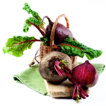 Two Fresh Raw Organic Beet Roots with Green Beet Tops and One Half in Wicker Basket on Wooden Board and Napkin isolated on White background