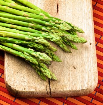 Arrangement of Fresh Raw Asparagus Sprouts on Wooden Cutting Board closeup on Orange Strawmat background