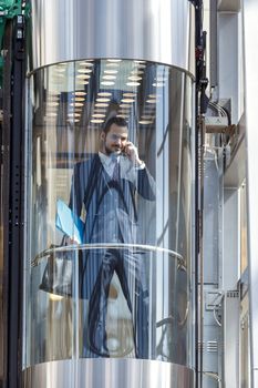 Businessman in modern glass elevator talking by the phone and holding papers