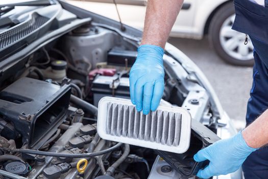 Auto mechanic wearing protective work gloves holds a dirty air filter over a car engine. Internal combustion engine air filter.