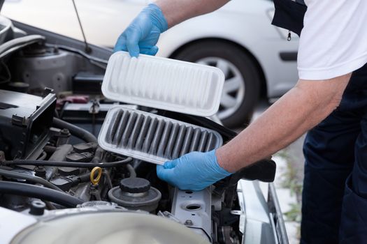 Auto mechanic wearing protective work gloves holds dirty and clean air filters over a car engine. Internal combustion engine air filters.