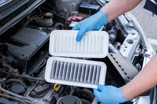 Auto mechanic wearing protective work gloves holds dirty and clean air filters over a car engine. Internal combustion engine air filters.