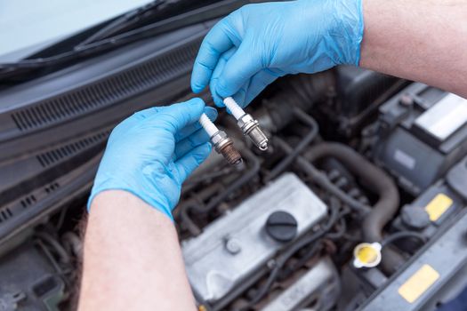 Auto mechanic wearing protective work gloves holds old and new spark plugs over a car engine