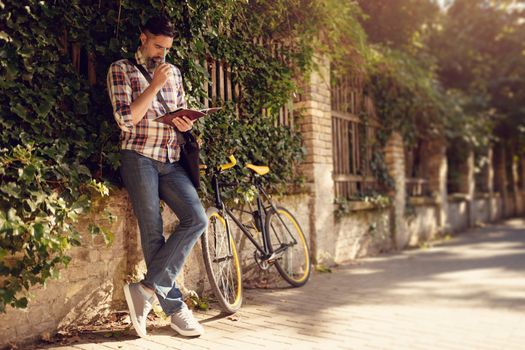 Pensive casual businessman taking his work to greener spaces. He is standing against the wall next to bike.