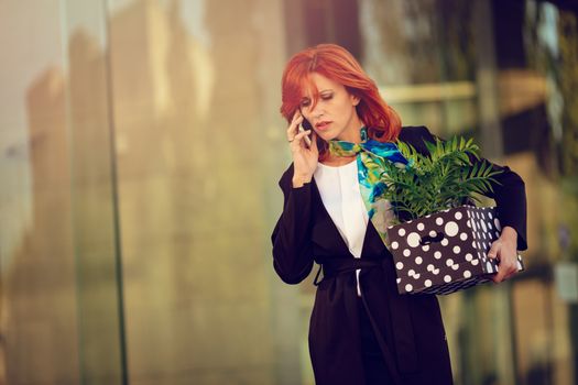 Serious pensive businesswoman walking in office district and talking on smartphone with a box full of her personal belongings from the office just after she got fired. 