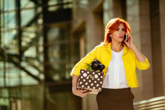 Serious pensive businesswoman walking in office district and talking on smartphone with a box full of her personal belongings from the office just after she got fired. 