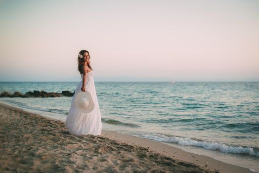 Beautiful young smiling woman in white dress walking on the beach and holding white summer hat.