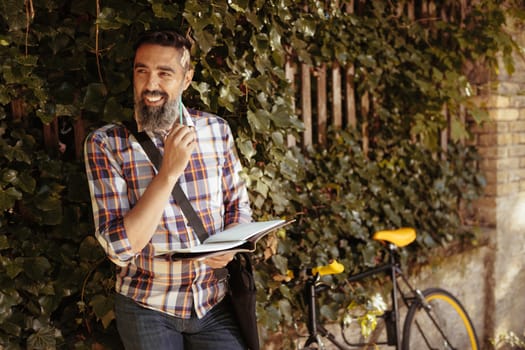 Smiling casual businessman working outdoor. He is standing against the wall next to bike.