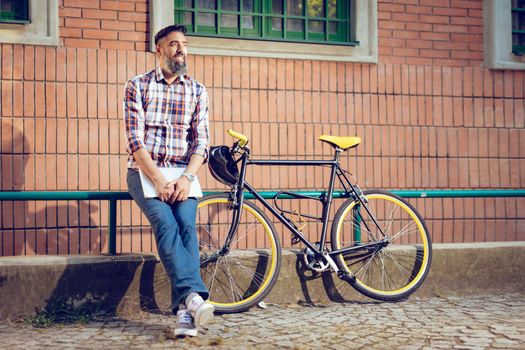 Smiling casual businessman having a break. He is standing in front of the building next to bike and holding laptop.