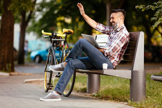 Smiling casual businessman waving Hello on a coffee break. He is sitting on a bench and working at laptop, next the bench rests a bike.