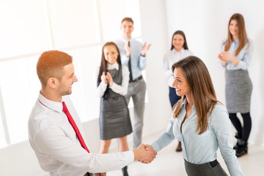 Successful young businesswoman and businessman shaking hands over a deal in front their business team with hands raised celebrating.