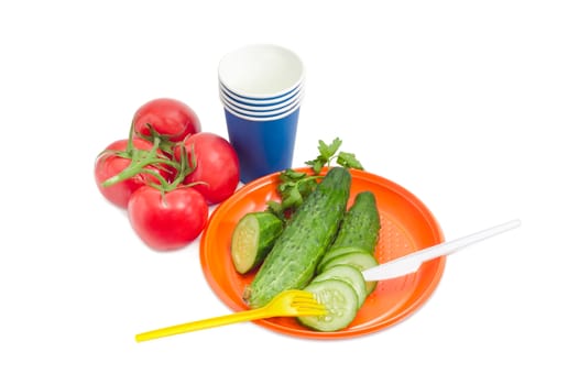 Disposable plastic plate, whole and sliced cucumber with fork and knife on it, cluster of the tomatoes, blue paper disposable cups beside on a light background
