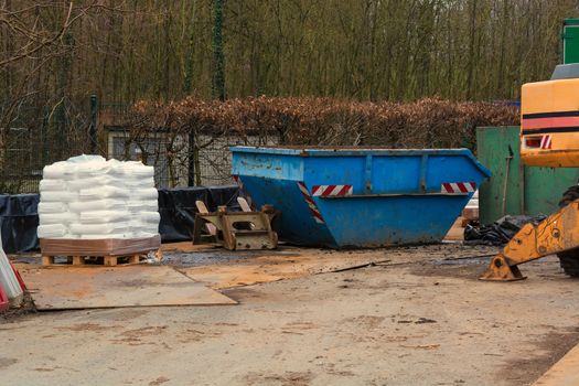 Blue container an excavator and pallet with white bags on a construction site in the forest.