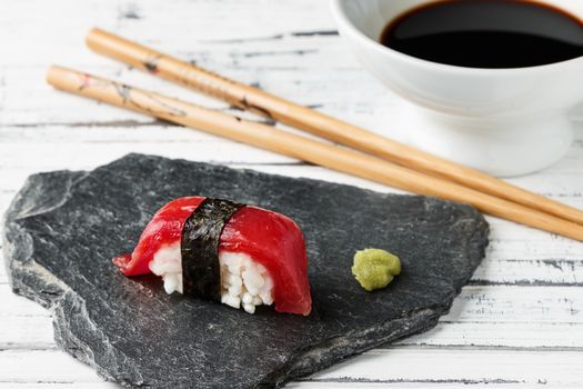 Red tuna Nigiri with Nori seaweed and wasabi paste on slate stone. Chopsticks and bowl with soy sauce in the background on old white wood. Raw fish in traditional Japanese sushi style. Horizontal image.