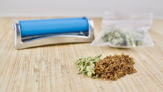 Blue rolling machine, bag with weed, and marijuana on wooden desk.