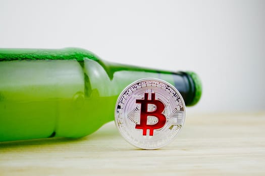 Cryptocurrency physical silver bitcoin coin with red sign near green beer bottle.