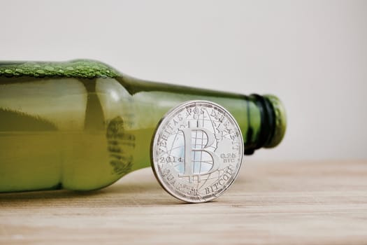 Digital currency physical silver bitcoin coin near green beer bottle on the wooden board.