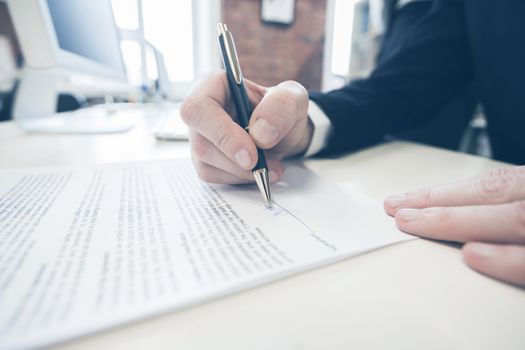 Businessman sitting at office desk signing a contract with shallow focus on signature.
