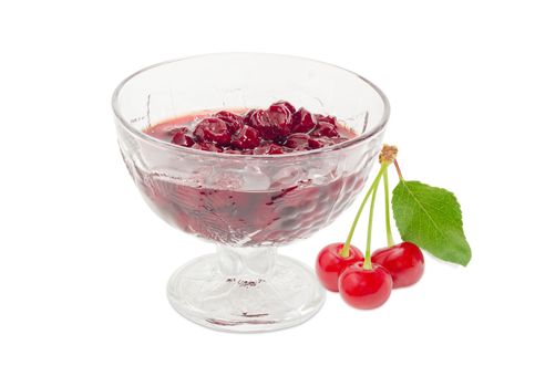 Cherry jam with whole fruits in the glass dessert stem bowl and three fresh cherries with the stalks and leaf beside on a light background
