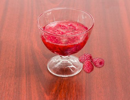 Raspberry jam in the glass dessert stem bowl and several fresh berries beside on a red wooden table
