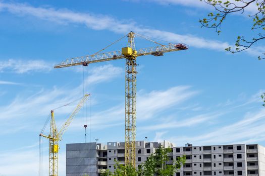 Two different tower cranes with latticed booms on the background of the upper part of a multi-story residential building under construction, tree branches and sky
