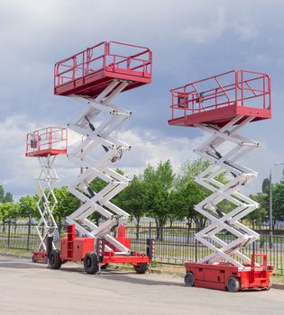 Three different red and white scissor wheeled lifts on an asphalt ground on the background of sky with clouds and trees along street
