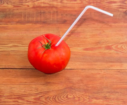 Big fresh ripe tomato with bendable drinking straw inserted into it on a surface of the old wooden planks
