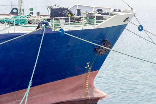 Bow of the old cargo ship with a blue starboard moored by several mooring ropes to the berth of the sea cargo port closeup
