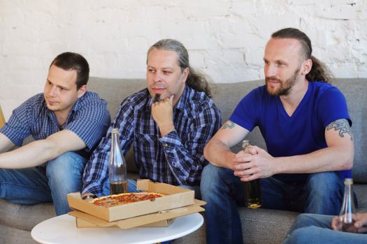 Group of men drinking beer, eating pizza, talking and smiling while resting at home on couch behind TV